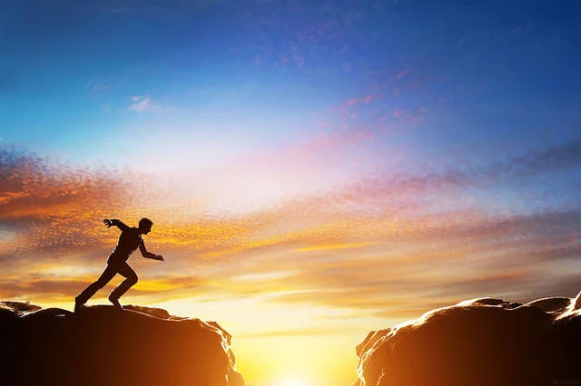 Silhouette of a person jumping across a gap between two rocky cliffs at sunset